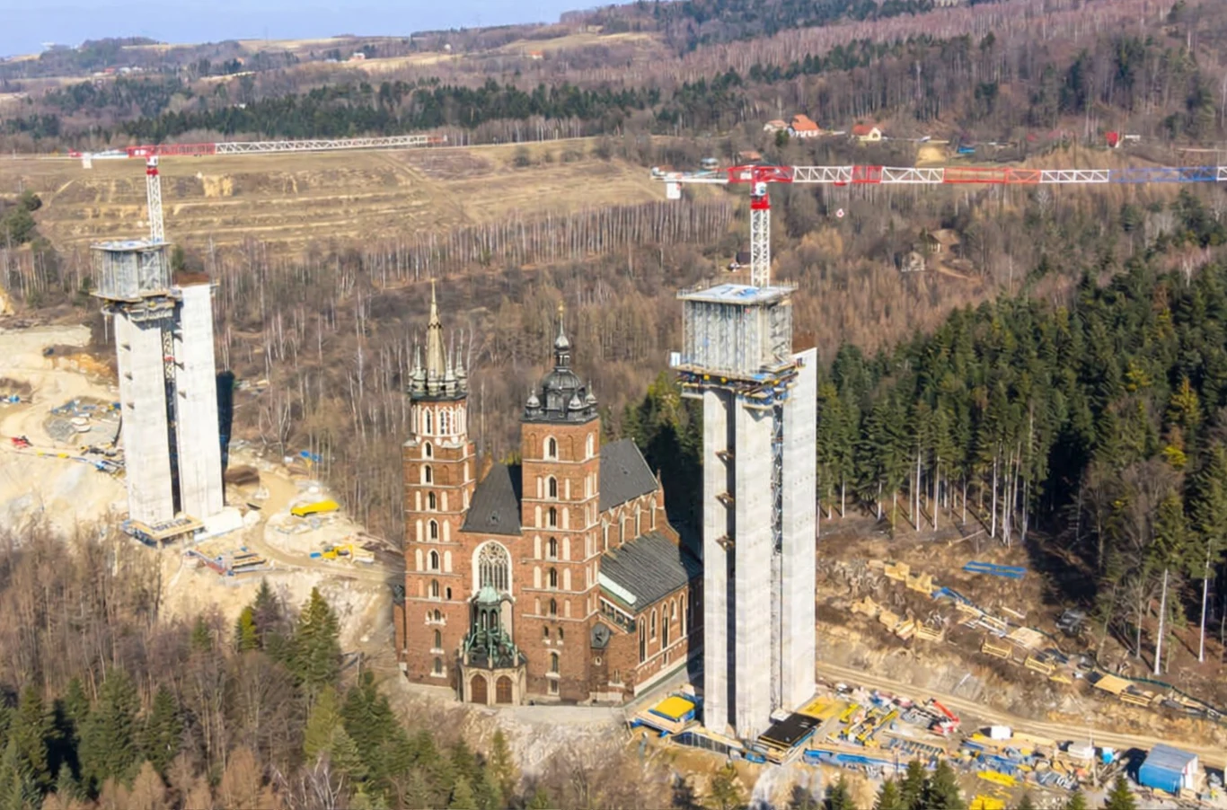 Ceglana świątynia z dwoma wysokimi wieżami otoczona przez las, po jej bokach dwa nowoczesne, betonowe pylony będące częścią konstrukcji mostu, widoczne dźwigi oraz maszyny budowlane, w tle rozległy krajobraz z polami i wzgórzami.