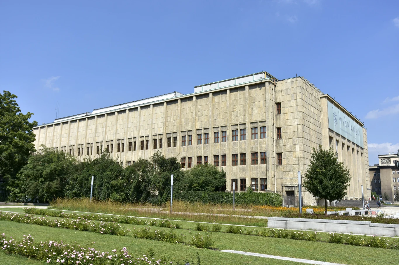 Monumentalny, prostokątny budynek z jasnego kamienia z regularnie rozmieszczonymi oknami, otoczony zadbaną zielenią i krzewami, na pierwszym planie widoczny jest murawa z alejkami i rabatami kwiatów.