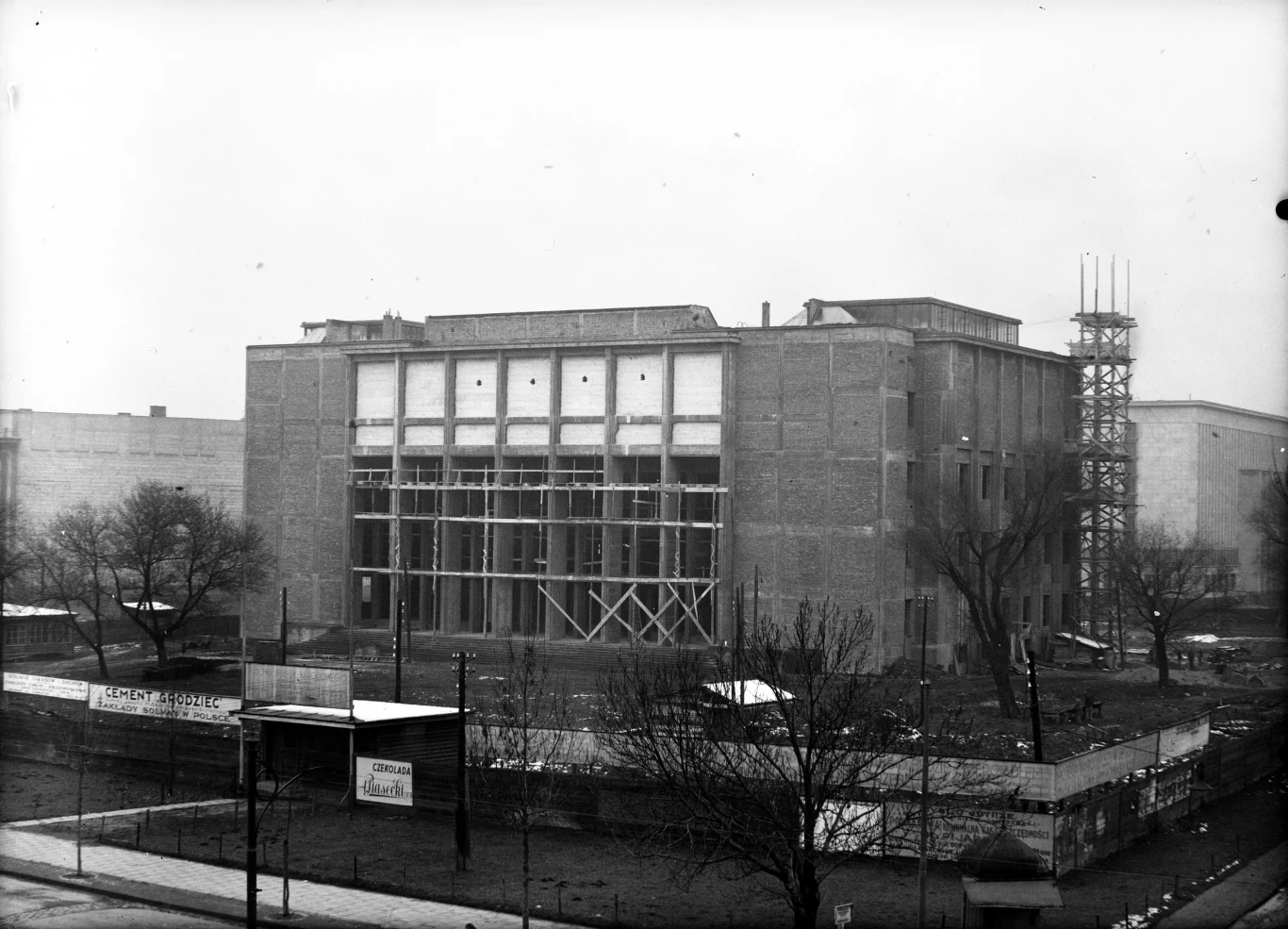 Duży, murowany budynek w trakcie budowy otoczony ogrodzeniem, z rusztowaniem po prawej stronie i niewykończoną fasadą, wokół drzewa bez liści oraz pusta ulica na pierwszym planie.