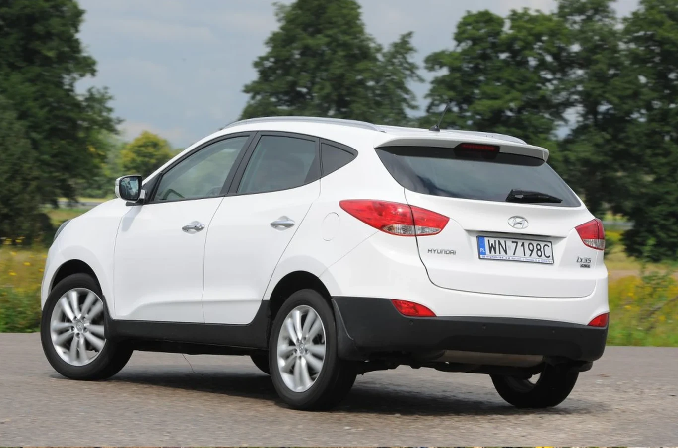 Biały samochód typu SUV marki Hyundai stoi na asfaltowej drodze, w tle widoczne wysokie drzewa na zielonej łące.