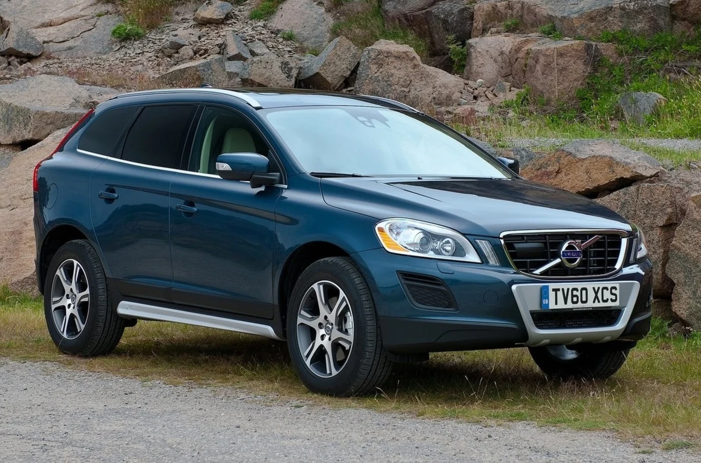 Granatowy SUV Volvo XC60 z jasnymi reflektorami i aluminiowymi felgami, zaparkowany na żwirowej nawierzchni w otoczeniu dużych kamieni.