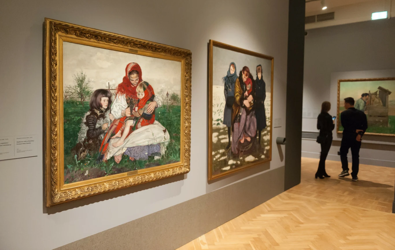 Wnętrze galerii sztuki z kilkoma dużymi obrazami przedstawiającymi grupy ludzi; na pierwszym planie obraz w złotej ramie ukazujący kobietę w czerwonej chuście z dziećmi siedzącymi na ławce, dalej para ogląda kolejny obraz na jasno oświetlonej ścianie.