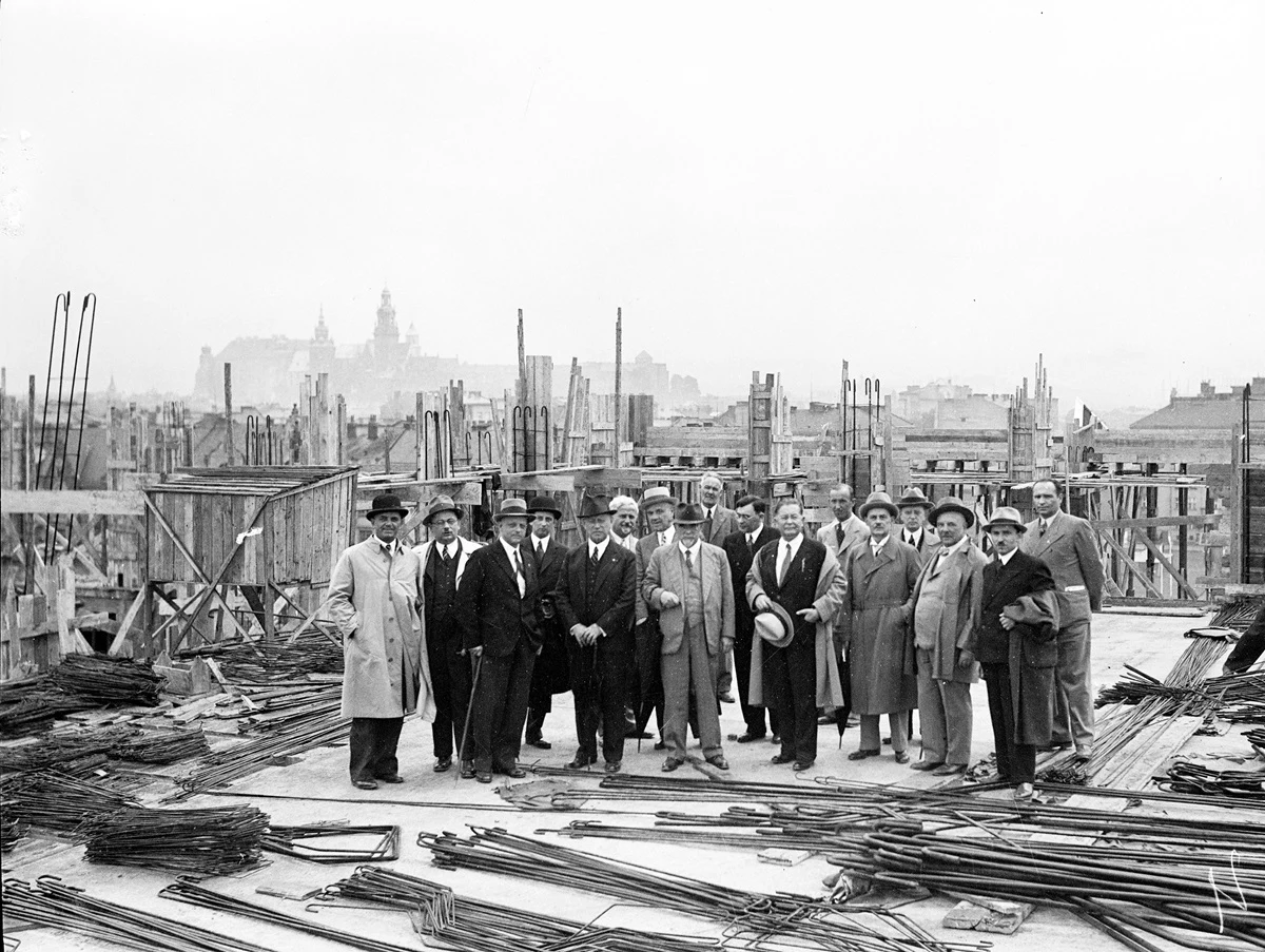 Grupa mężczyzn w garniturach i płaszczach stoi pośród stalowych prętów i drewnianych konstrukcji na placu budowy, w tle widoczne zarysy miasta z charakterystycznym zamkiem.