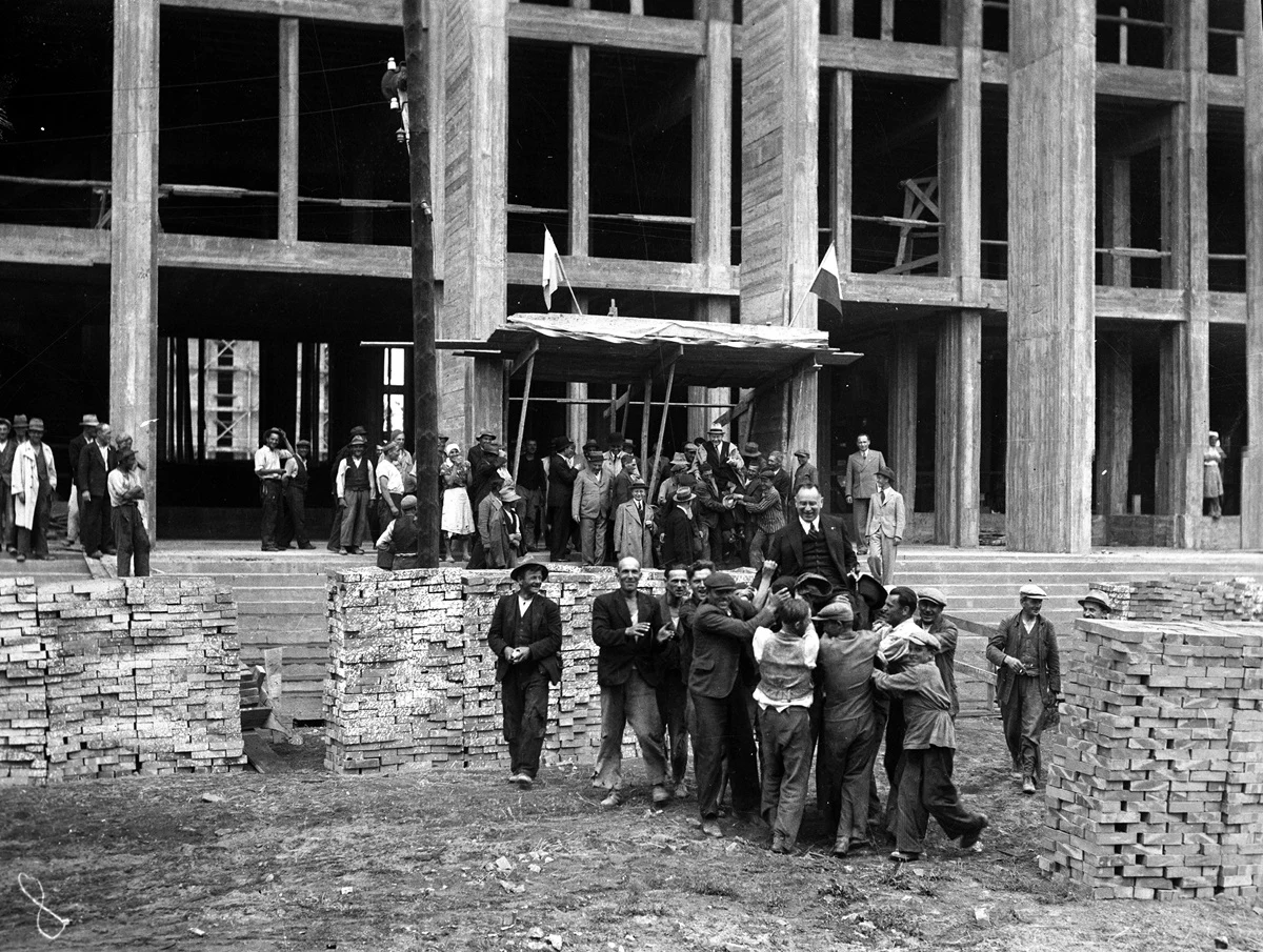 Grupa ludzi niesie mężczyznę na noszach na tle budującego się budynku z surowego betonu, wokół stoją inni obserwatorzy i cegły ułożone w stosy po obu stronach.