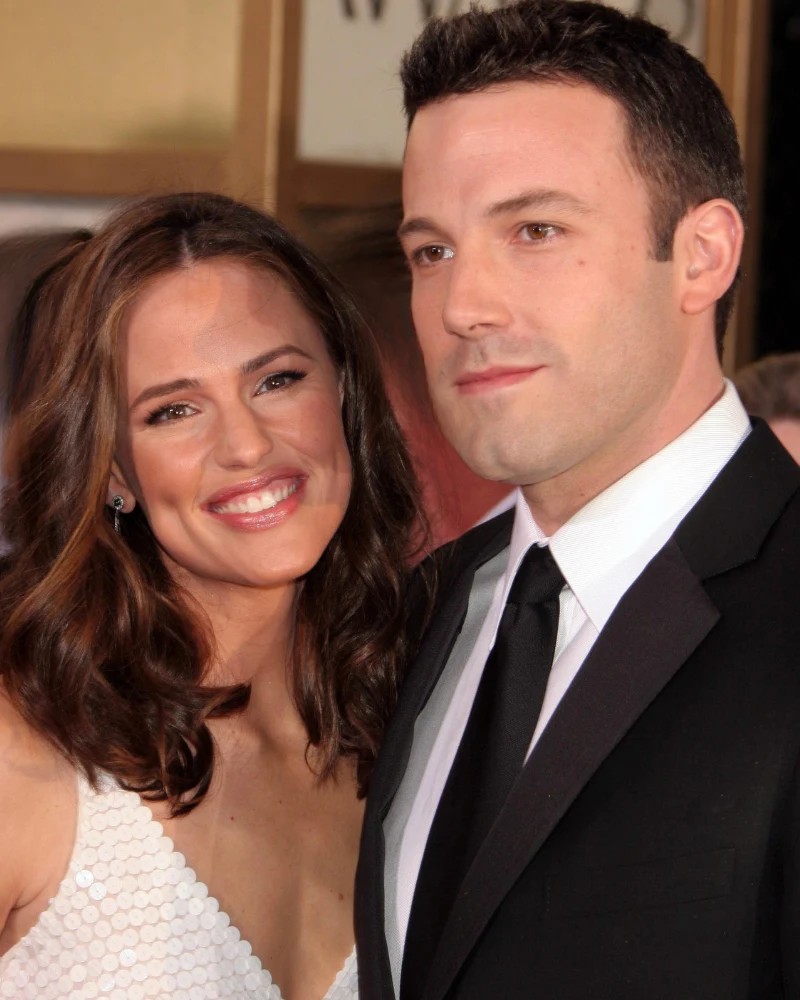 Kobieta z długimi, falowanymi włosami w białej sukience uśmiecha się, stojąc blisko mężczyzny w eleganckim czarnym garniturze i białej koszuli z krawatem; oboje patrzą w stronę obiektywu.