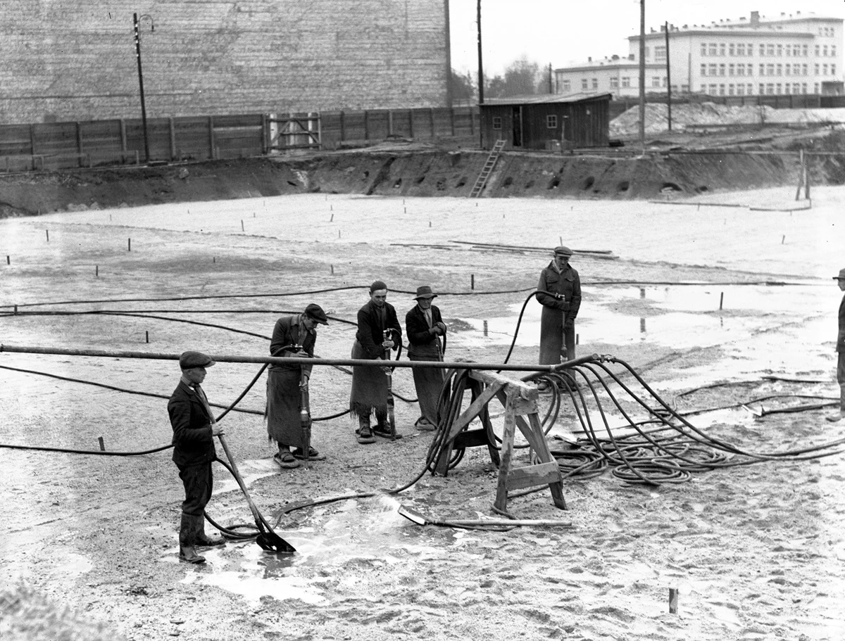 Grupa mężczyzn pracujących przy systemie węży i rur na zalanym terenie budowy, otoczonym niskim ogrodzeniem, w tle duży budynek przemysłowy oraz zabudowania miejskie.