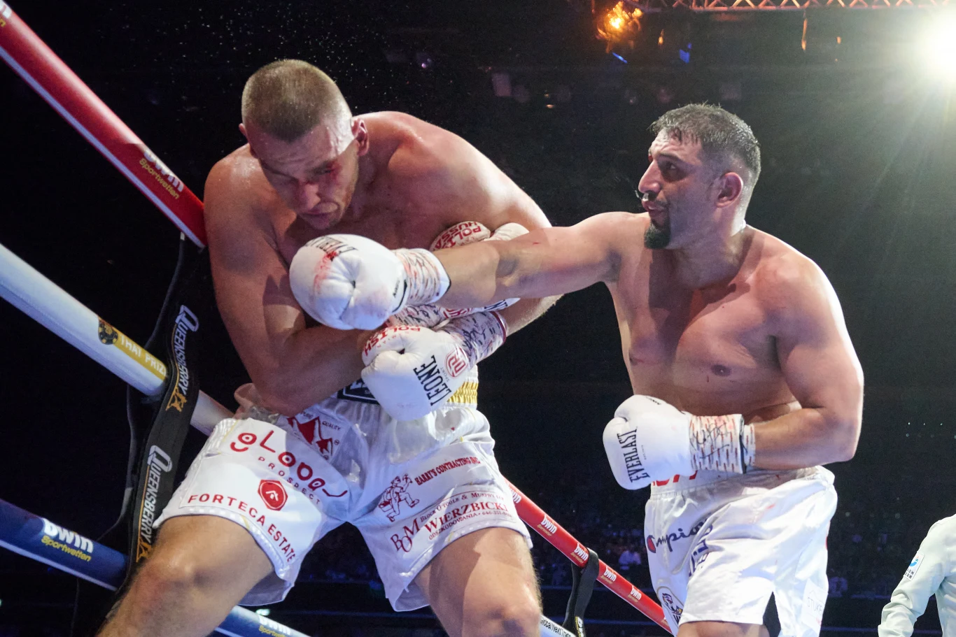 Dwóch bokserów w białych spodenkach podczas zaciętej walki, jeden z zawodników zadaje silny cios prawą ręką prosto w głowę przeciwnika, który osłania się rękami przy linach ringu.