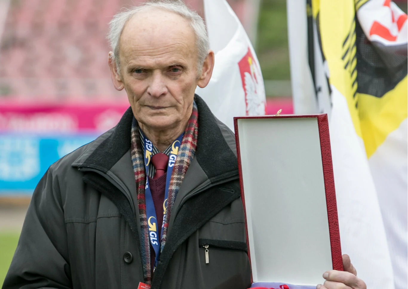 Starszy mężczyzna w ciemnej kurtce trzyma otwarte pudełko na medal lub odznaczenie, w tle widoczne flagi i rozmyte trybuny stadionu.