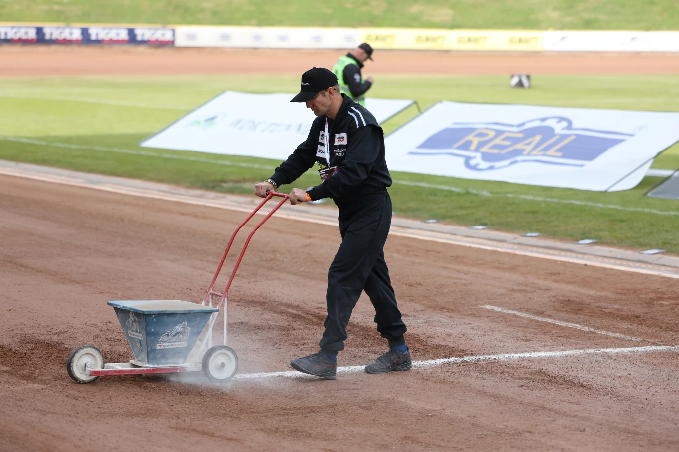 Mężczyzna w czarnym stroju sportowym oznacza linię kredą na torze żużlowym, korzystając z wózka do malowania linii.