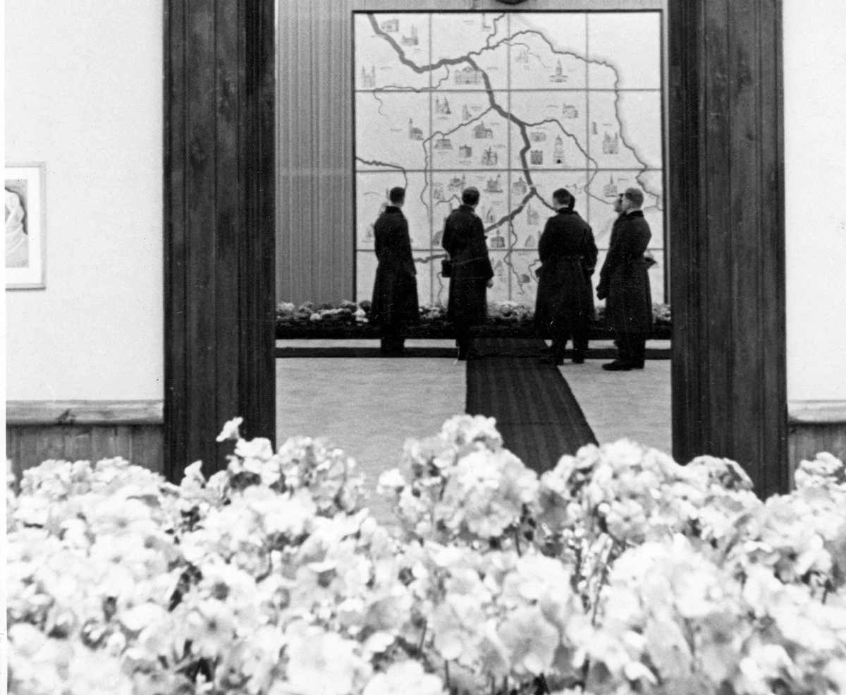 Pięciu mężczyzn w płaszczach stoi przed dużą mapą ścienną, analizując szczegóły, w pierwszym planie widoczne są kwiaty ozdobne, scena utrzymana w czarno-białej tonacji.