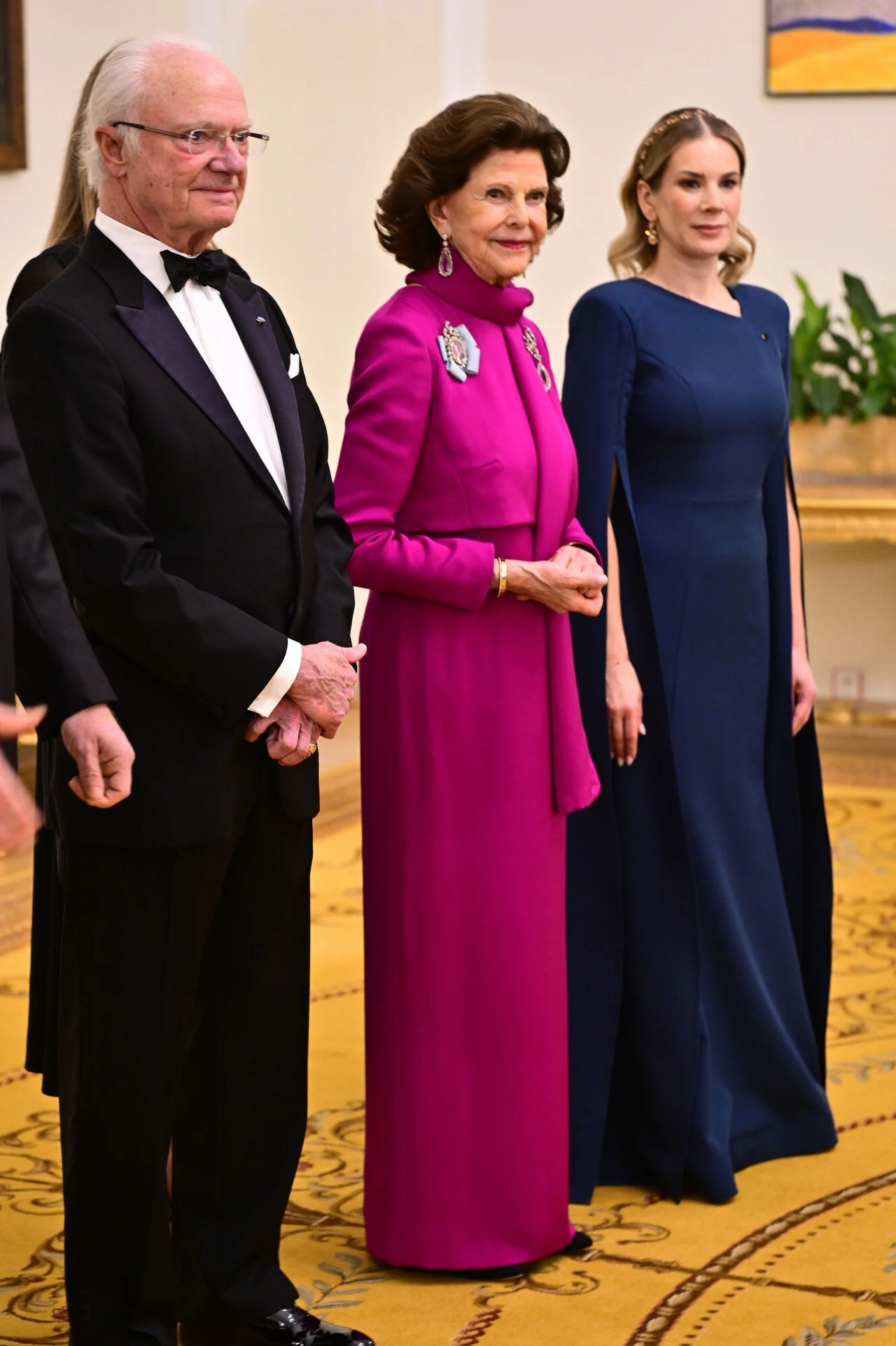 Trzy elegancko ubrane osoby stojące na żółtym dywanie w ozdobnej sali; mężczyzna w smokingu obok kobiety w długiej, fuksjowej sukni z broszką oraz kobiety w granatowej, długiej sukni z rozcięciami na rękawach.