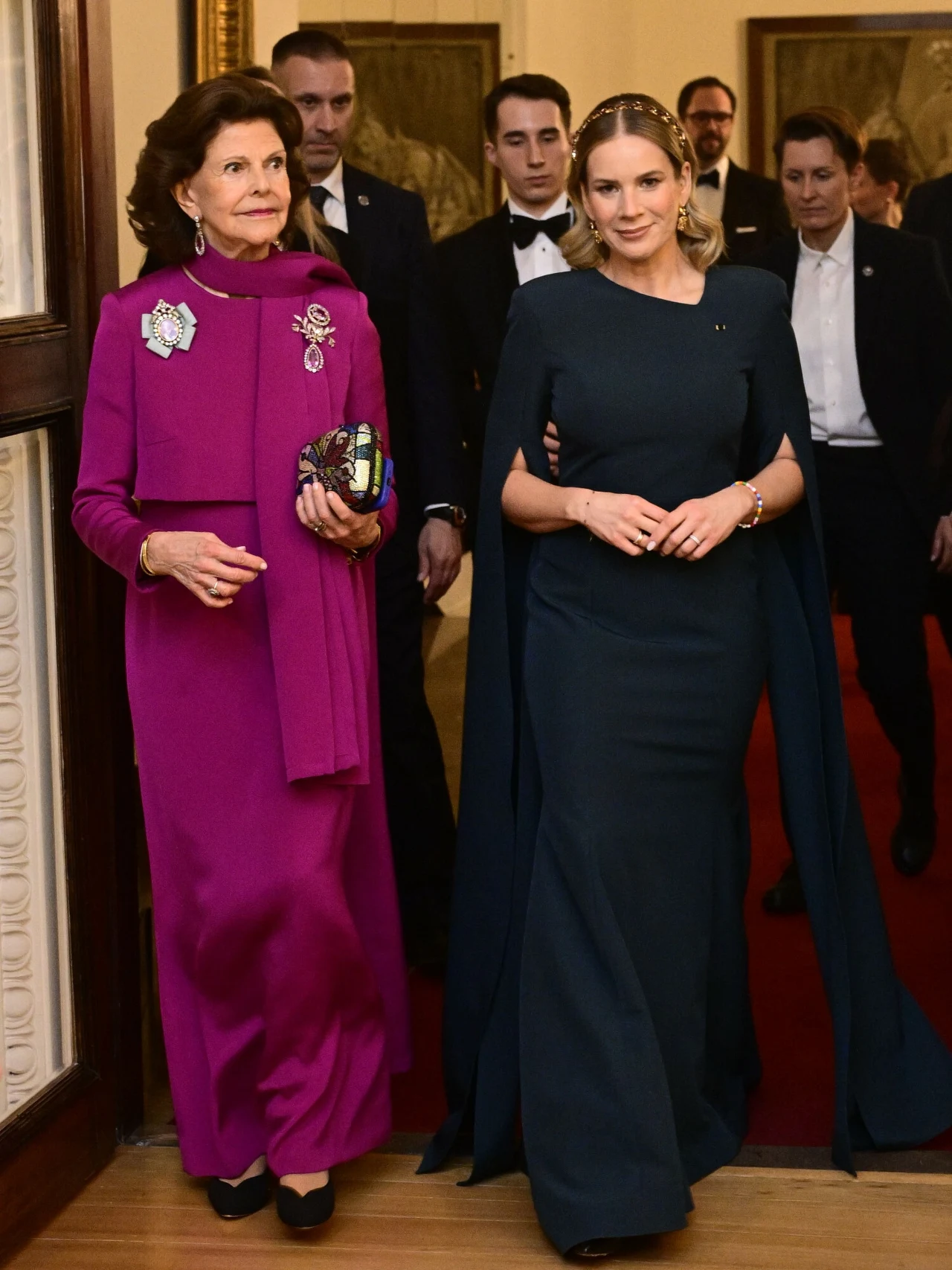 Dwie elegancko ubrane kobiety idą obok siebie, jedna w długiej fioletowej sukni z broszkami i zdobioną torebką, druga w ciemnozielonej sukni z długimi pelerynami, wokół nich kilku mężczyzn w smokingach, całość w otoczeniu pałacowych wnętrz i czerwonego...
