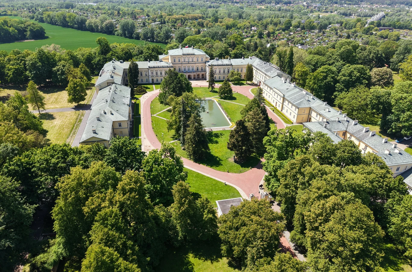 Rozległy pałac z symetrycznie ustawionymi skrzydłami otaczający dziedziniec z centralnym fontanną, otoczony gęstym parkiem i ścieżkami, widoczny z lotu ptaka.