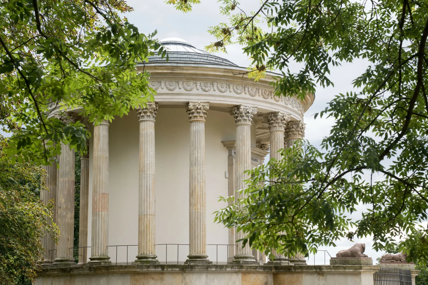 Klasyczna rotunda otoczona kolumnami w stylu antycznym, częściowo przesłonięta gałęziami drzew, z fragmentem ozdobnego gzymsu i rzeźbami lwów na podeście.