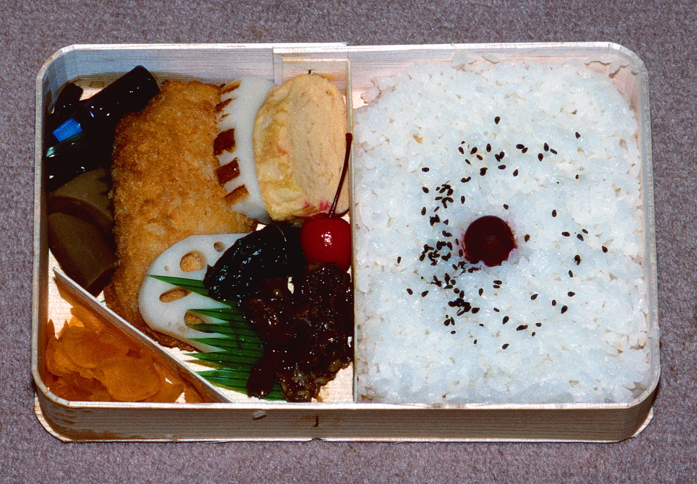 Bento z porcją białego ryżu posypanego czarnym sezamem i umeboshi oraz dodatkami: panierowanym kotletem, plasterkiem tamagoyaki, marynowanymi warzywami, kawałkiem ryby, lotosem i wiśnią.