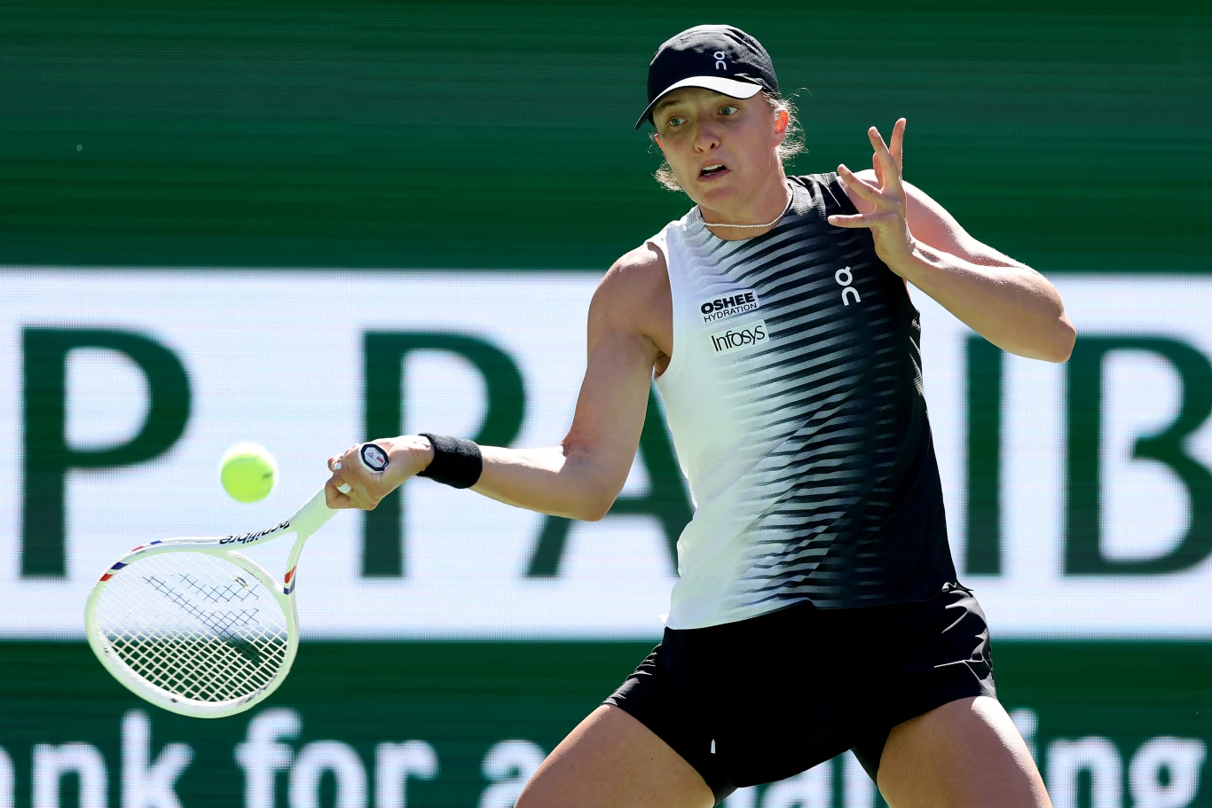 Tenisistka w czarno-białym stroju wykonuje forehand, skupiona na piłce lecącej tuż przed rakietą, na tle zielonego kortu z dużym logo sponsora.