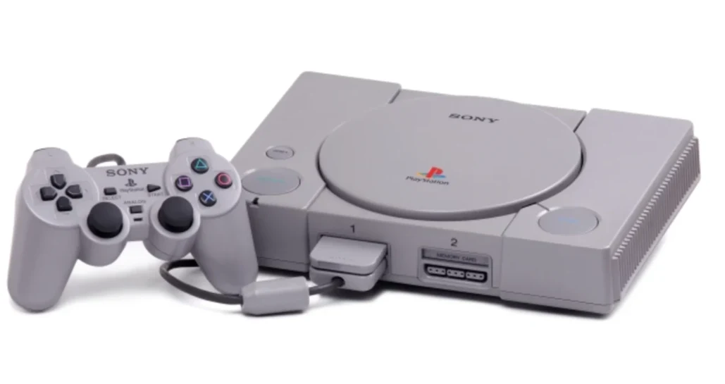 Szara konsola Sony PlayStation wraz z podłączonym kontrolerem DualShock, widoczne logo PlayStation na przednim panelu oraz przyciski funkcyjne urządzenia.