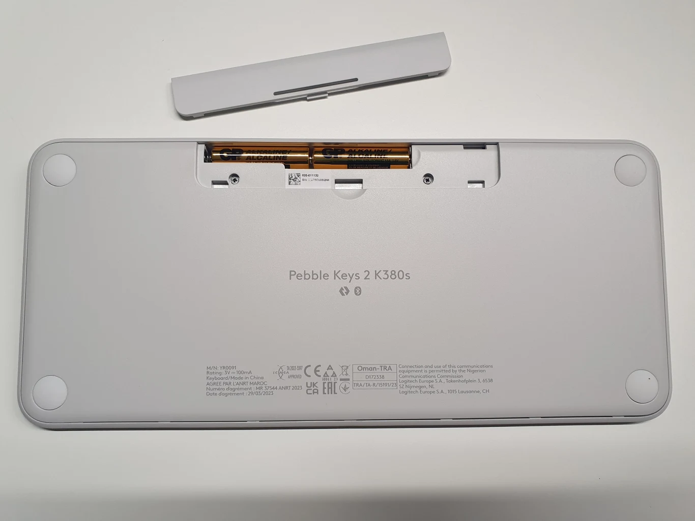 Biała klawiatura bezprzewodowa Pebble Keys 2 K380s odwrócona spodem do góry, z otwartą komorą na baterie oraz zdjętą pokrywą leżącą obok.