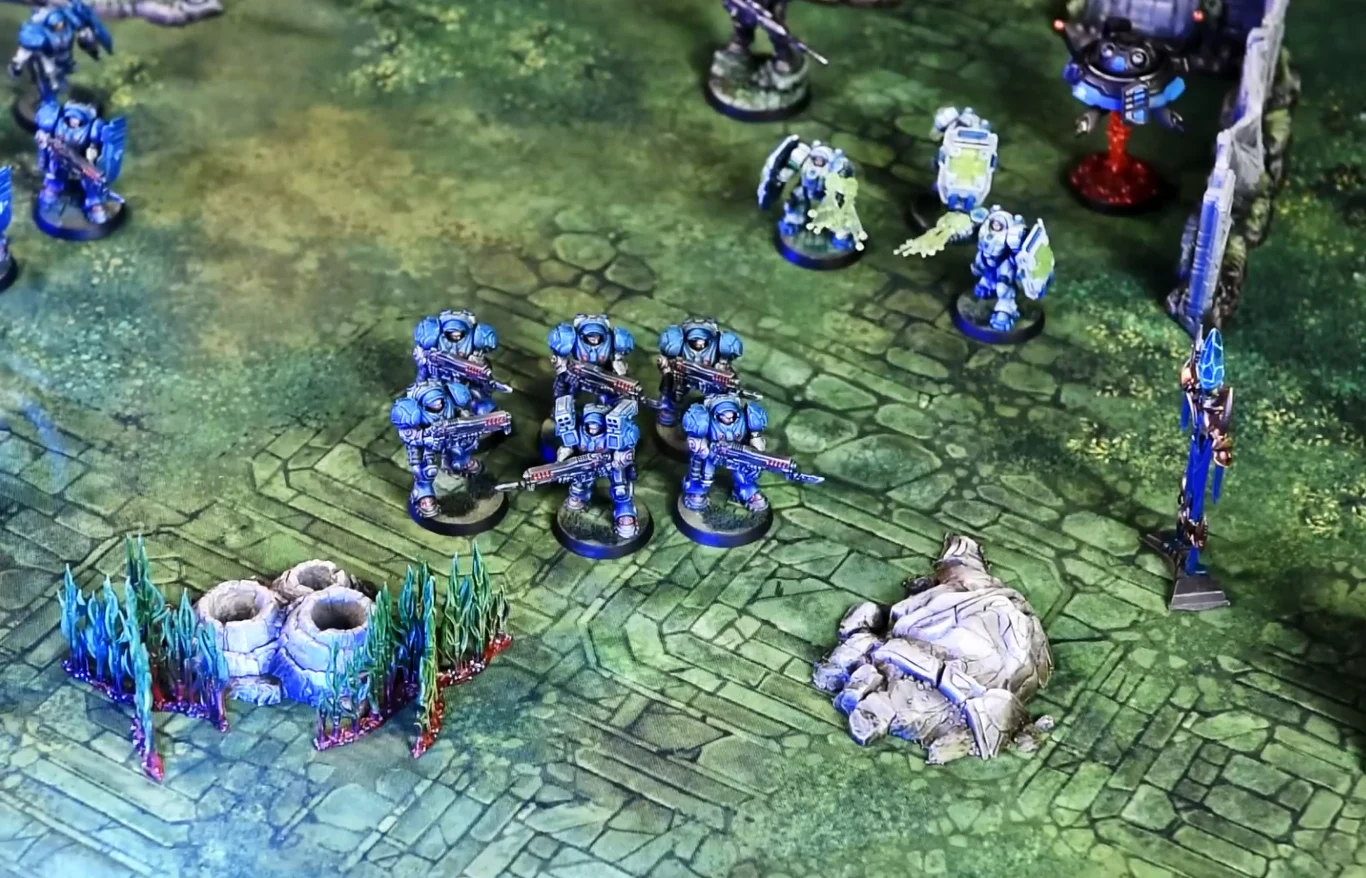 Cztery figurki żołnierzy StarCraft w niebieskich zbrojach na planszy z ruinami i elementami terenu.