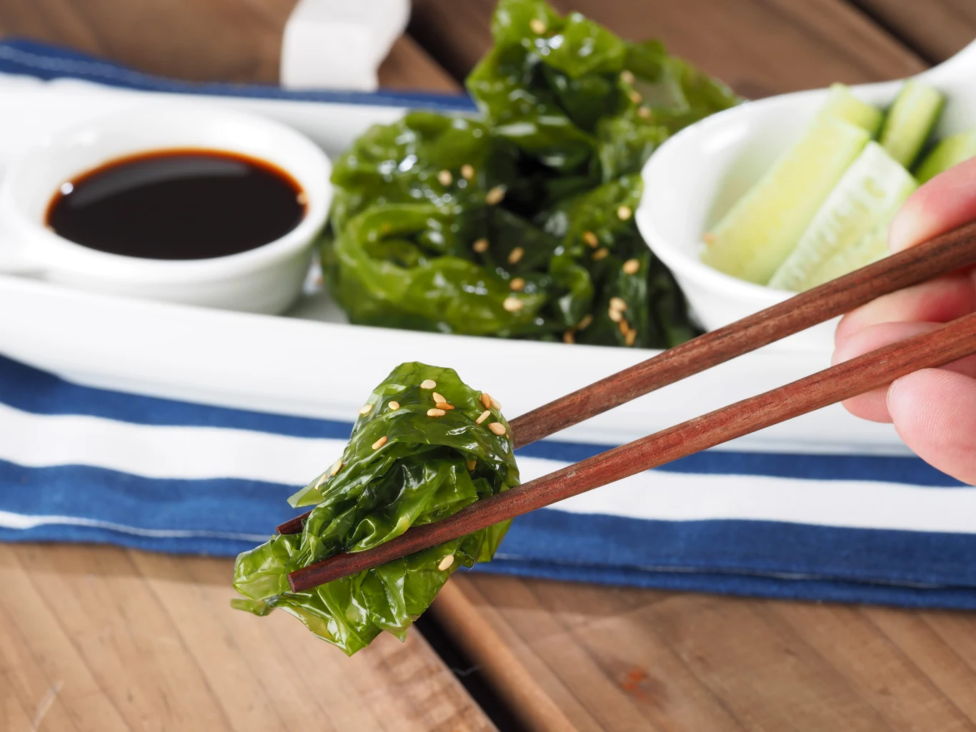Porcja sałatki z alg morskich posypana sezamem trzymana pałeczkami, w tle białe naczynie z sosem sojowym oraz kawałkami ogórka.