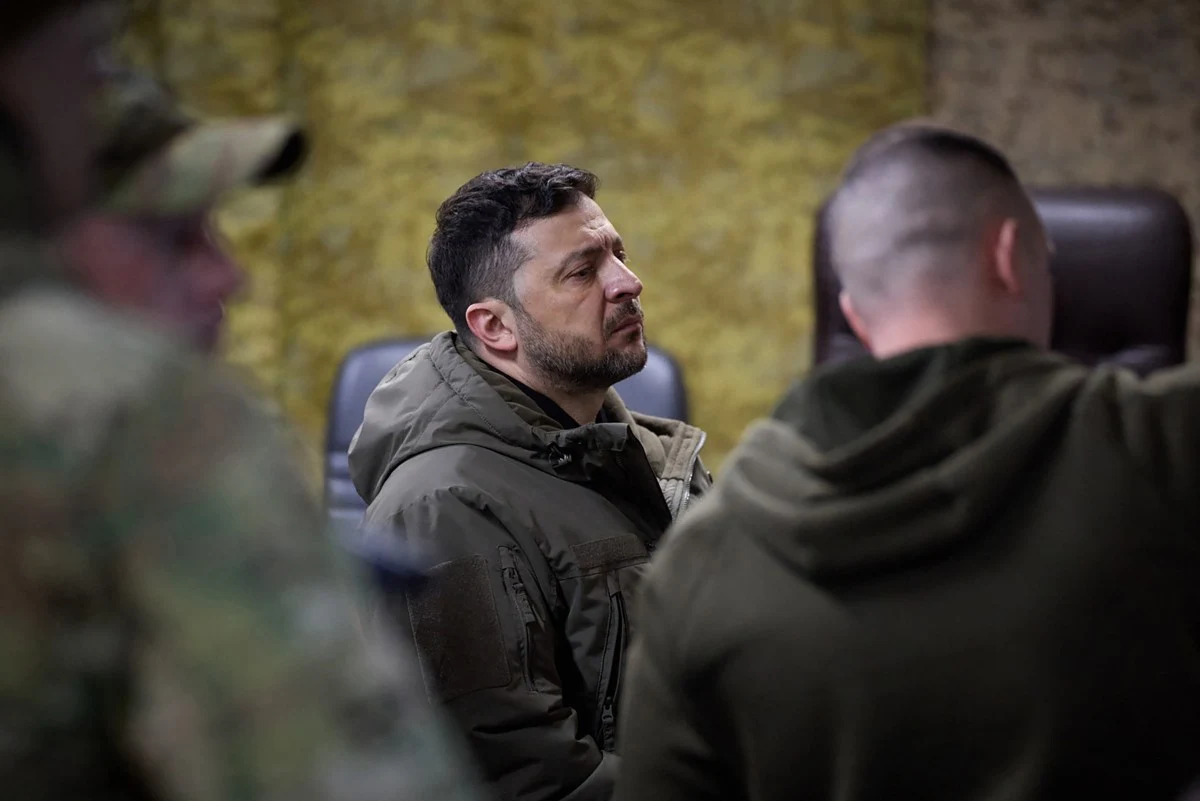 Mężczyzna w ciemnej kurtce skupiony na rozmowie z dwoma mężczyznami w wojskowych ubraniach na tle żółtej ściany, siedząc na krześle biurowym.