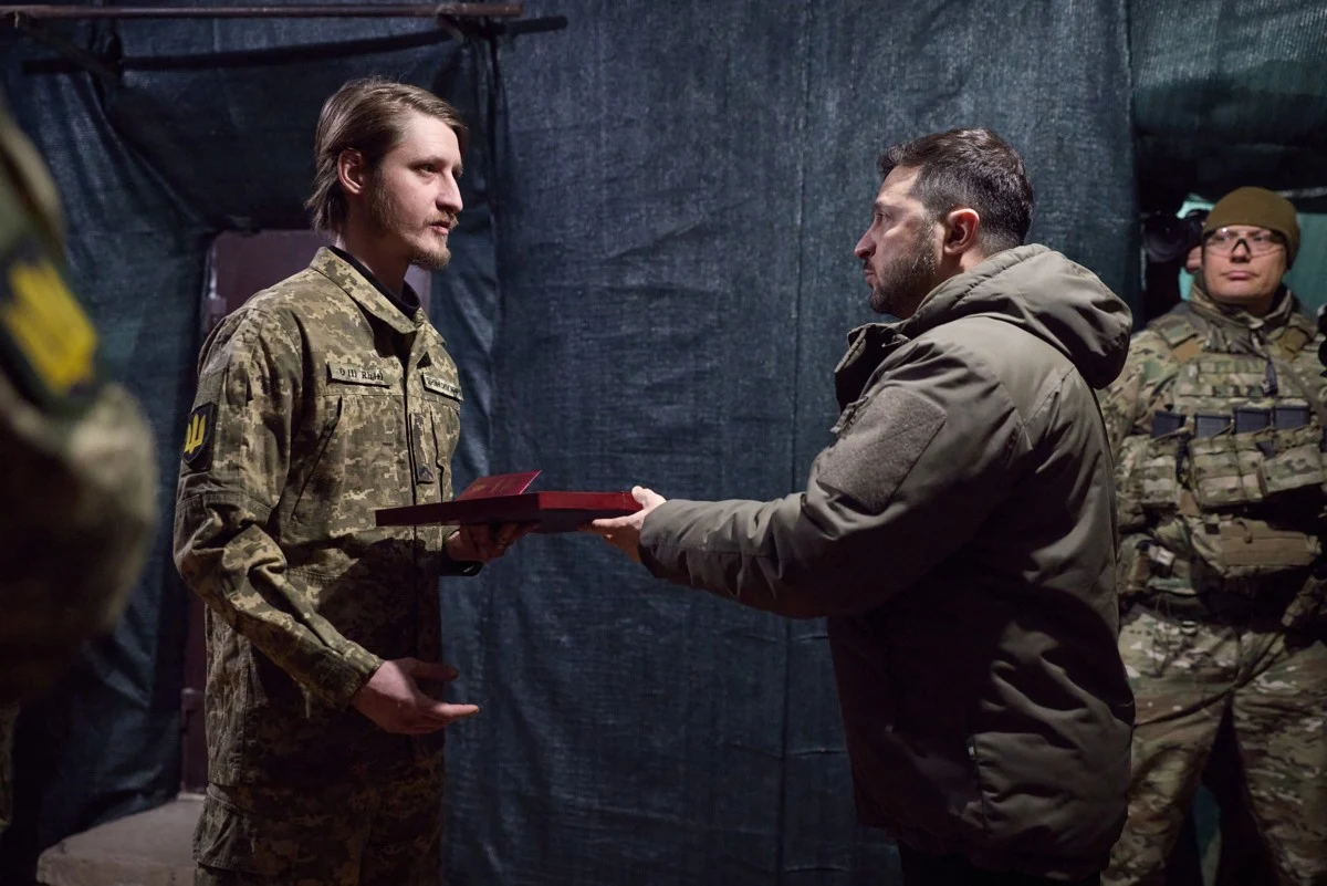 Dowódca w wojskowym mundurze wręcza odznaczenie żołnierzowi stojącemu naprzeciwko niego, w tle obecni są inni wojskowi obserwujący sytuację.