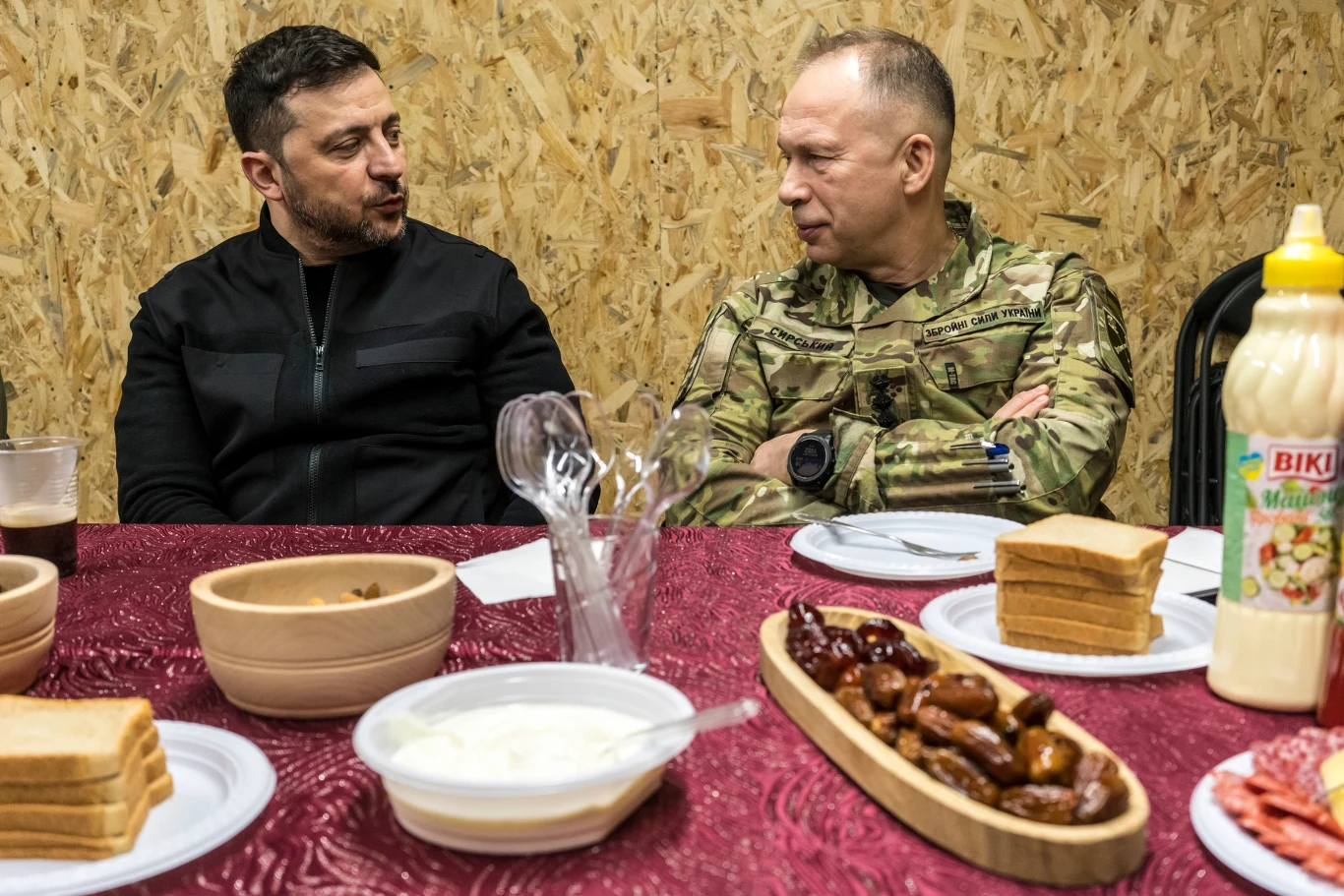 Dwóch mężczyzn, jeden w czarnym ubraniu, drugi w wojskowym mundurze, rozmawia przy stole nakrytym czerwonym obrusem, na którym ustawiono talerze z chlebem, kiełbaskami, plasterkami wędlin, sosami oraz plastikowymi miskami z dodatkami.