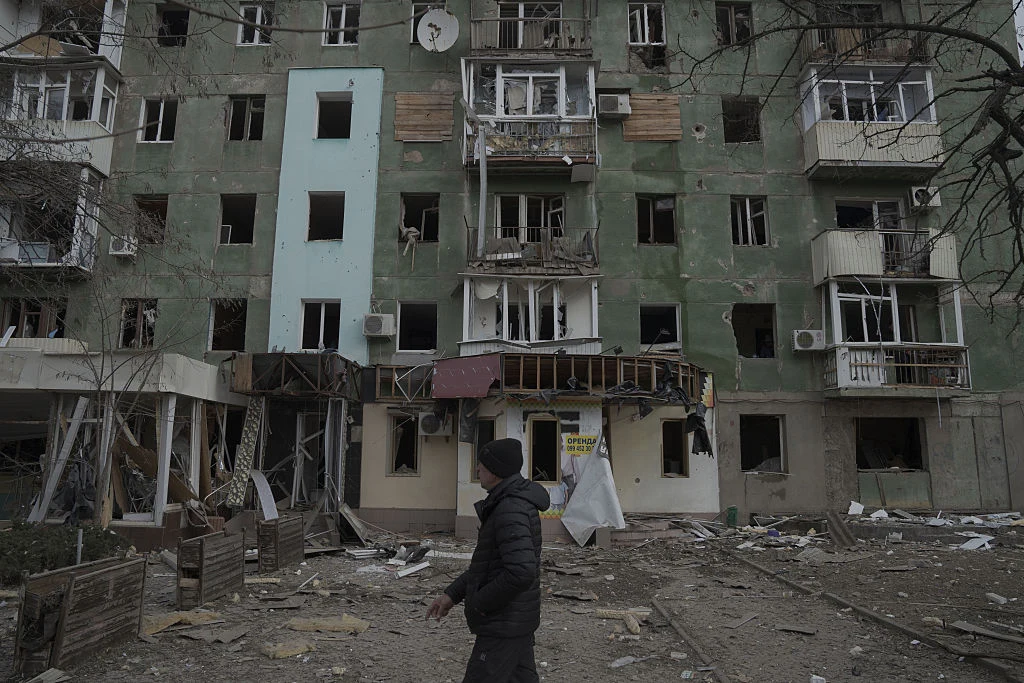 Zniszczona fasada wielopiętrowego bloku mieszkalnego z powybijanymi szybami i uszkodzonymi balkonami, gruzy rozrzucone przed budynkiem, na pierwszym planie samotna sylwetka przechodnia.