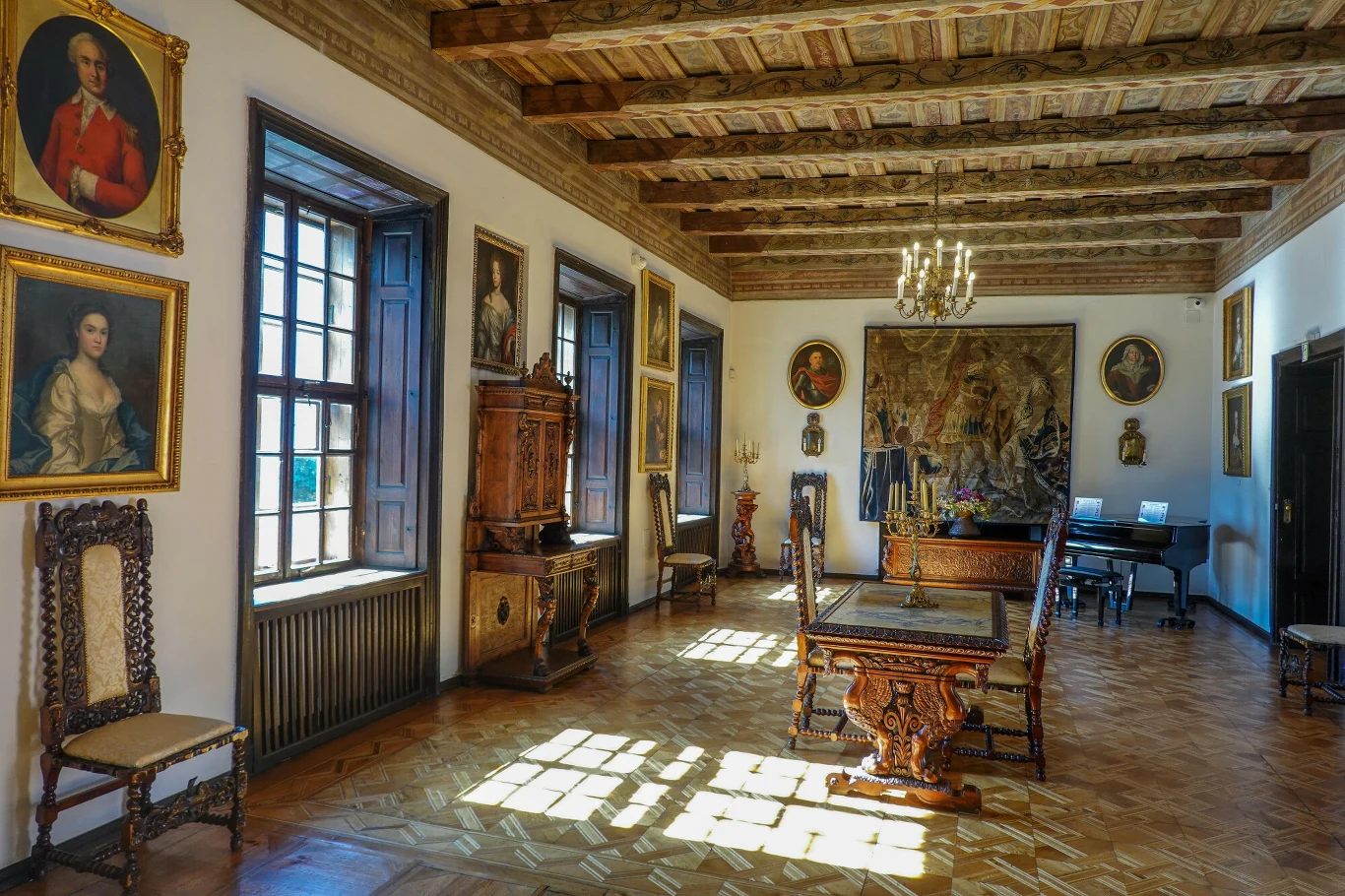 Zabytkowe wnętrze z drewnianym stropem, dużymi oknami wpuszczającymi naturalne światło, rzeźbionymi krzesłami i stołami o bogatych zdobieniach, licznymi portretami w złotych ramach na ścianach oraz gobelinem jako centralnym elementem dekoracyjnym.