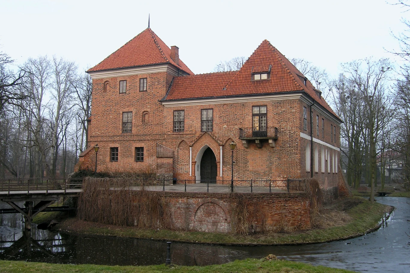 Ceglany zamek otoczony fosą, z mostem prowadzącym do głównego wejścia oraz licznymi oknami i dwuspadowym dachem pokrytym czerwoną dachówką, w tle bezlistne drzewa.