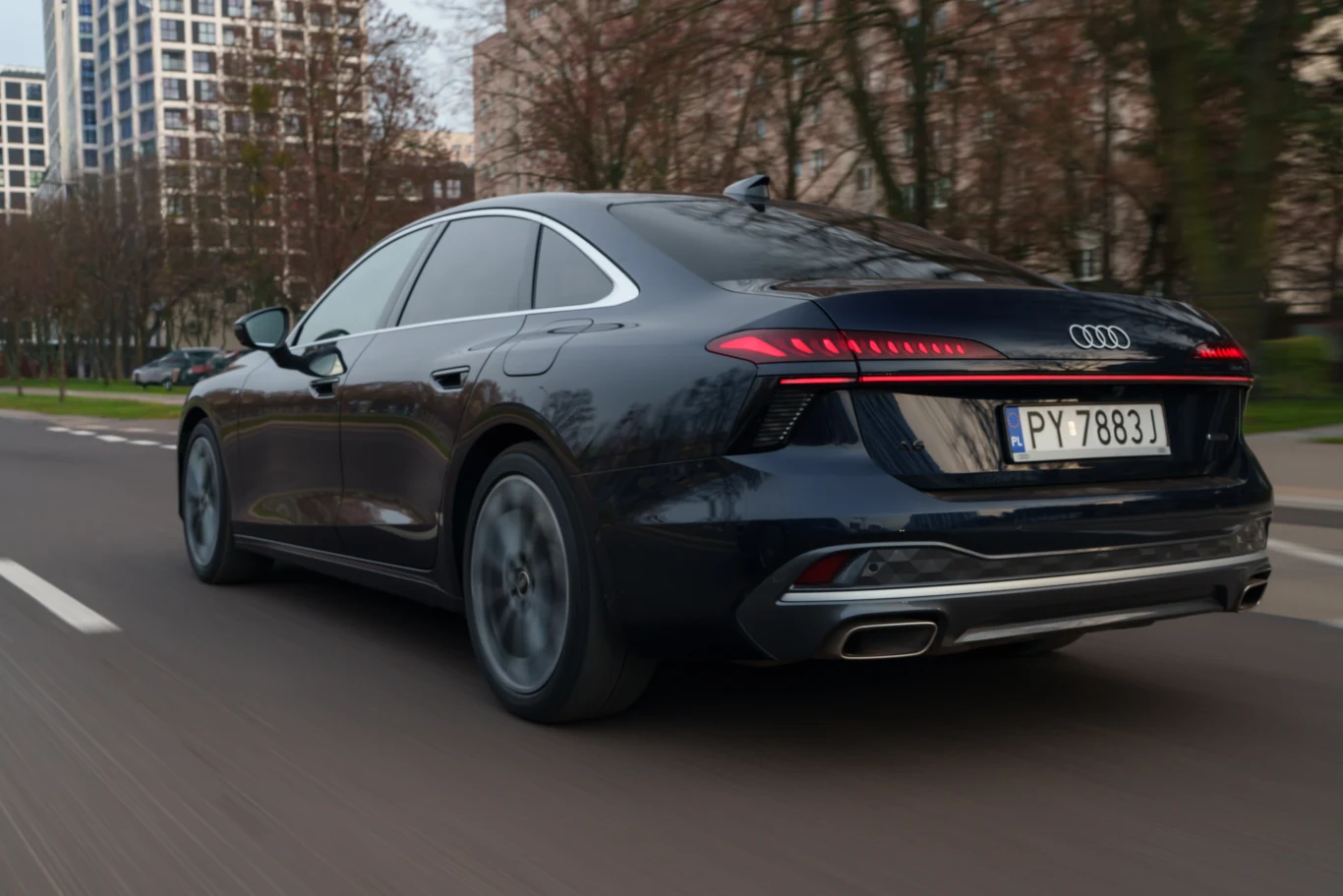Czarny sedan marki Audi poruszający się po miejskiej ulicy, charakterystyczne tylne światła LED i nowoczesna sylwetka pojazdu podkreślają jego dynamiczny styl.