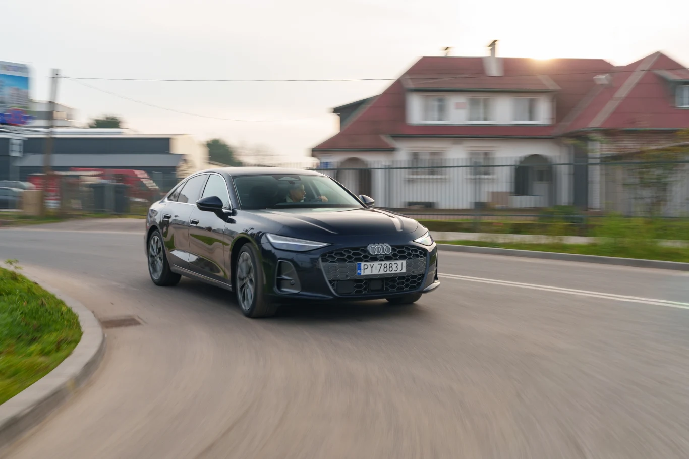 Czarny samochód marki Audi porusza się po zakręcie ulicy w osiedlu domów jednorodzinnych, w tle widoczny nowoczesny budynek z dwuspadowym dachem.