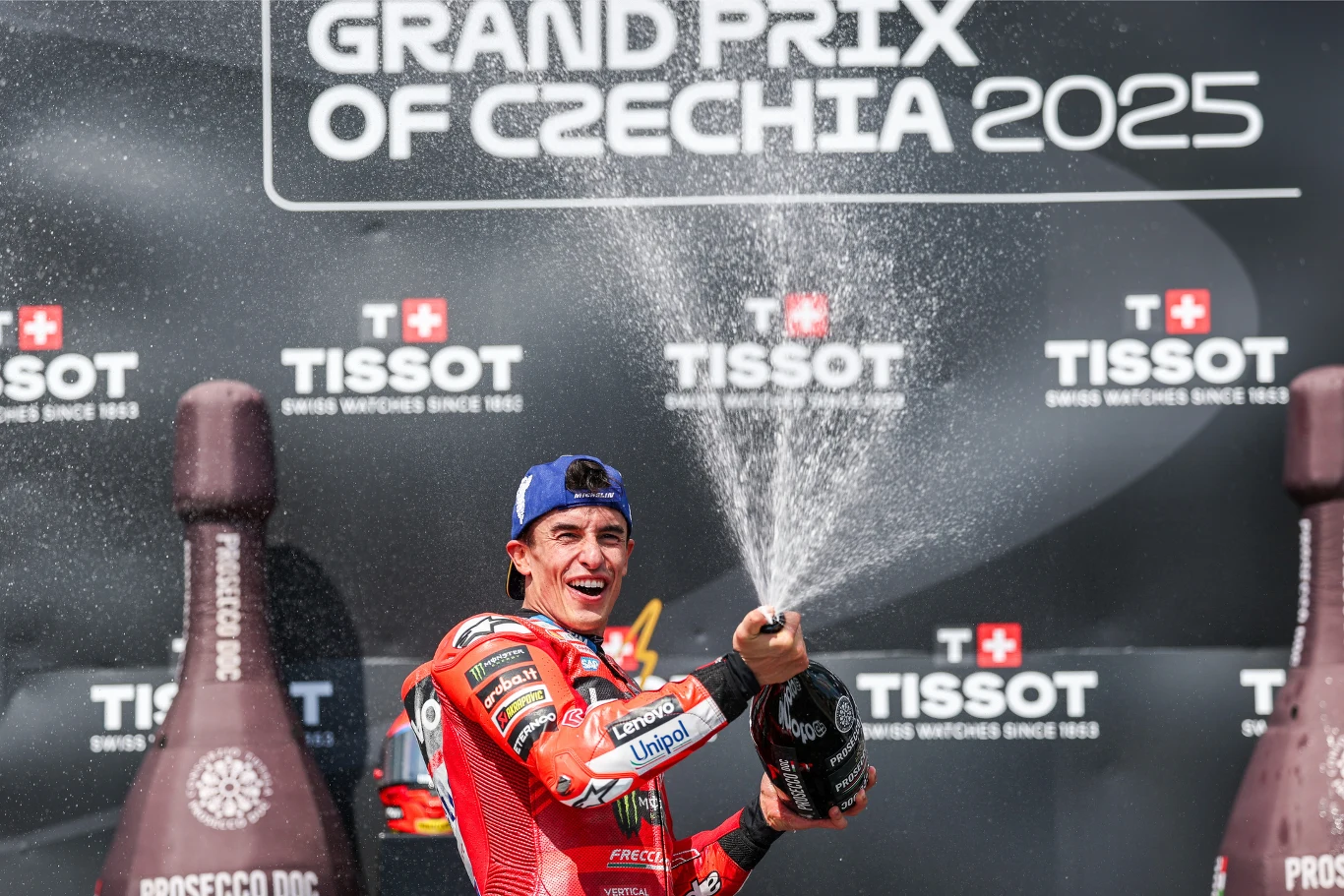 Zwycięzca wyścigu motocyklowego w czerwonym kombinezonie świętuje na podium, tryskając szampanem, w tle logo Grand Prix Czech oraz reklamy sponsora Tissot.