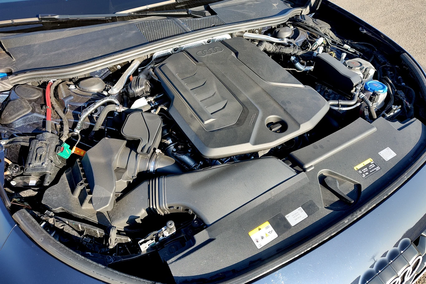 Silnik samochodowy z licznymi komponentami, przewodami i elementami mechanicznymi, widoczne logo Audi na osłonie silnika, dobrze uporządkowana komora silnika z plastikowymi osłonami i widocznym zbiornikiem płynu chłodniczego po prawej stronie.