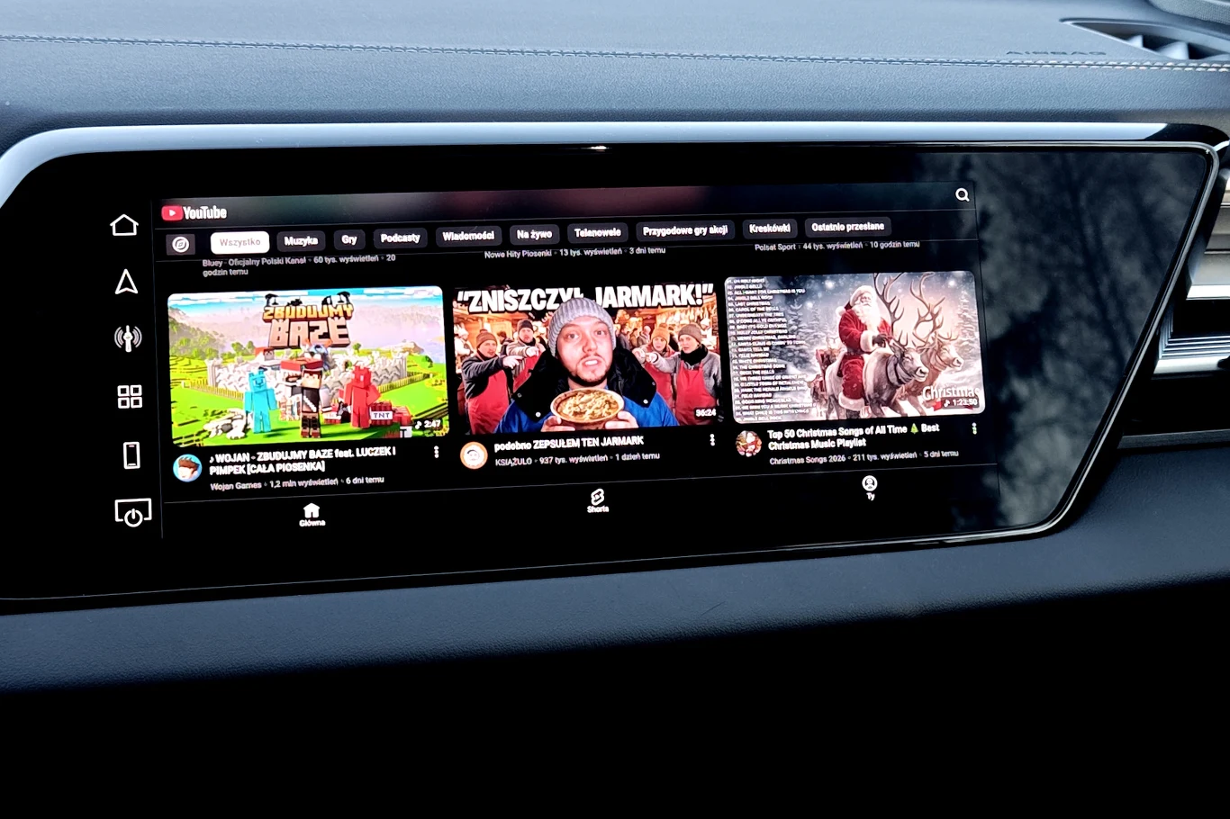 Ekran dotykowy samochodu wyświetlający stronę główną YouTube z trzema sugerowanymi filmami oraz ikonami nawigacji po lewej stronie panelu.
