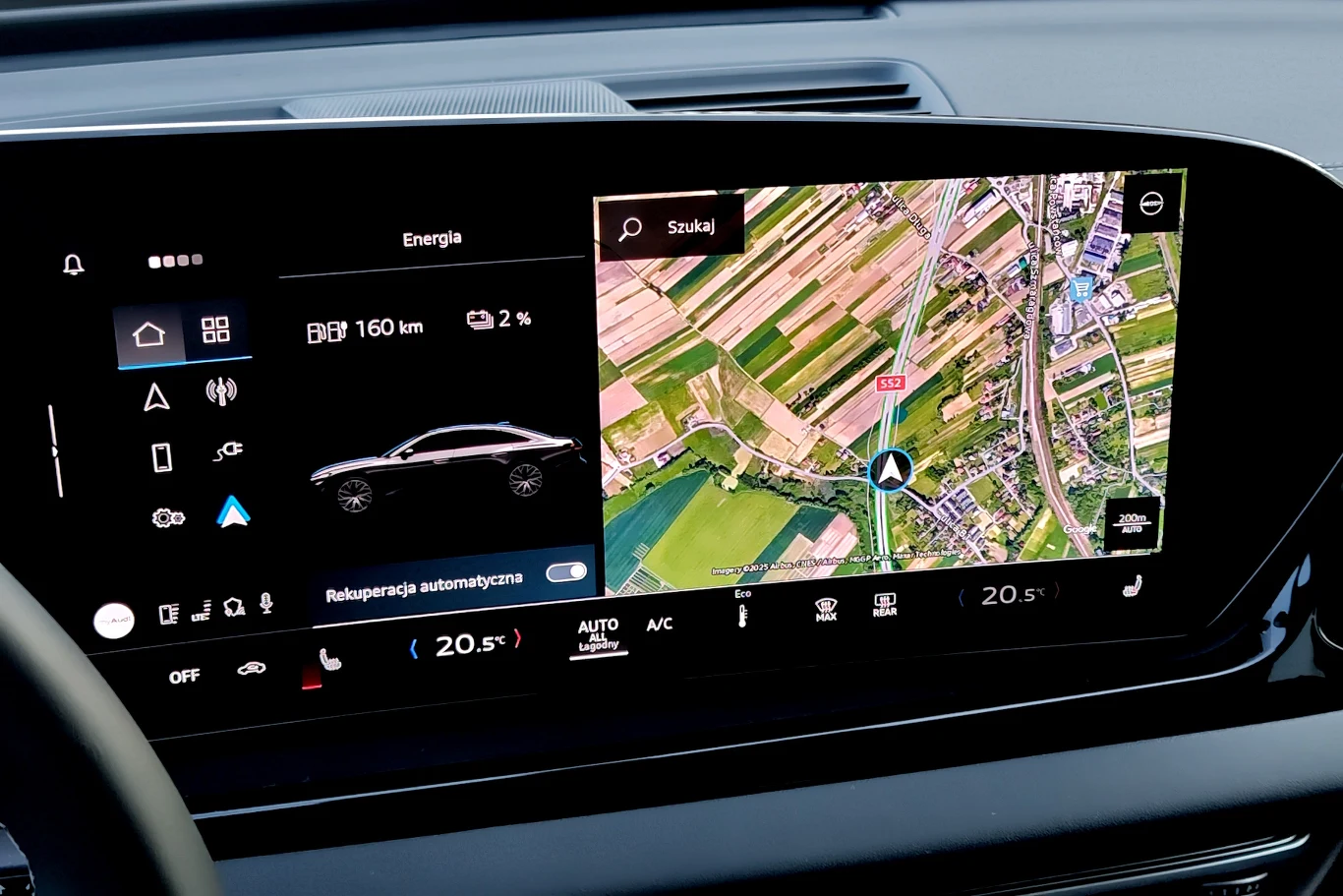 Cyfrowy wyświetlacz samochodowy z interfejsem pokazującym stan baterii, zasięg pojazdu elektrycznego oraz mapę nawigacji z widokiem satelitarnym i wyraźnie zaznaczoną bieżącą lokalizacją samochodu na tle pól uprawnych.