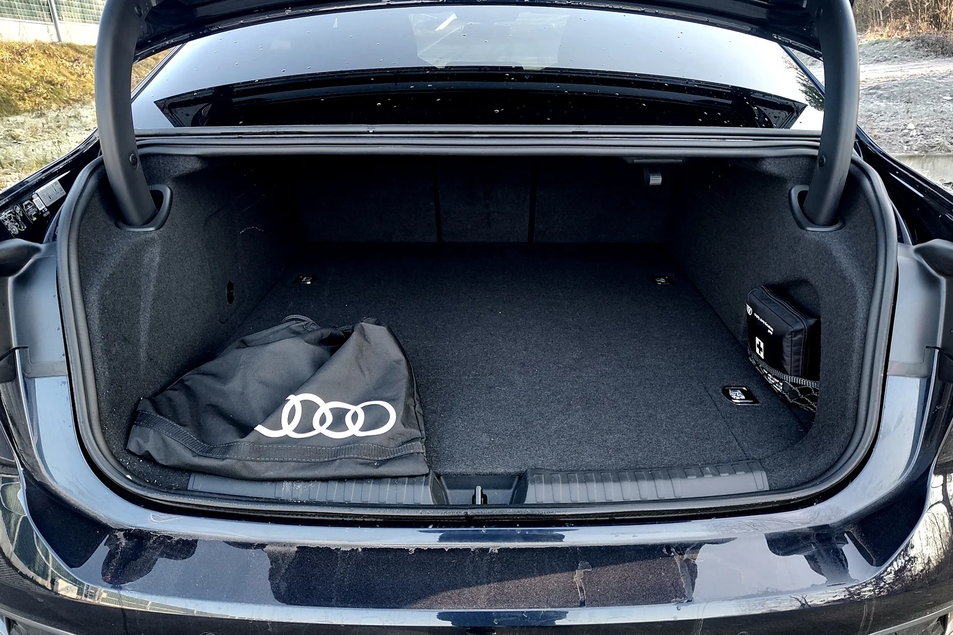 Otwarty bagażnik samochodu marki Audi z dużą przestrzenią ładunkową, po lewej stronie widoczna czarna torba z logo Audi, po prawej niewielkie czarne pudełko przymocowane do ściany bagażnika.
