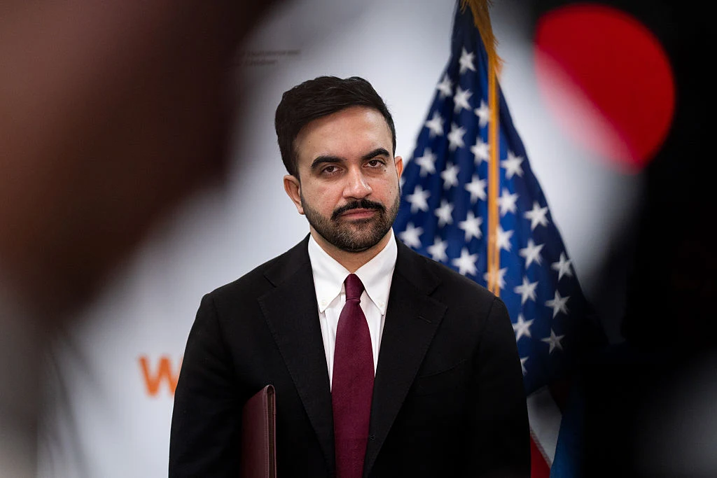 Mężczyzna w ciemnym garniturze i bordowym krawacie stoi na tle flagi Stanów Zjednoczonych, patrząc poważnie w stronę aparatu.