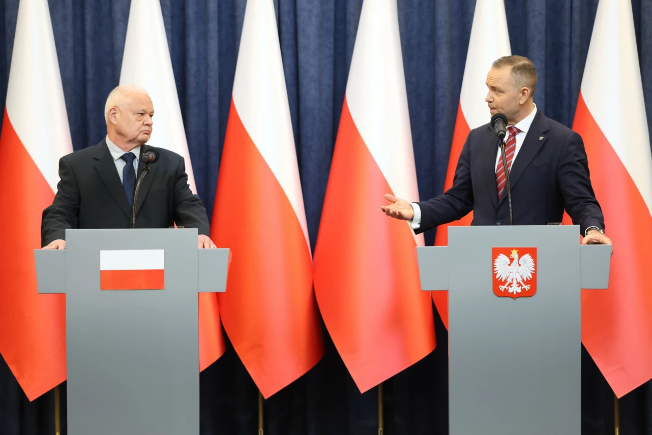Dwóch mężczyzn w garniturach stoi za mównicami z symbolami Polski na tle kilku polskich flag, jeden z nich trzyma mikrofon i gestykuluje w kierunku rozmówcy.