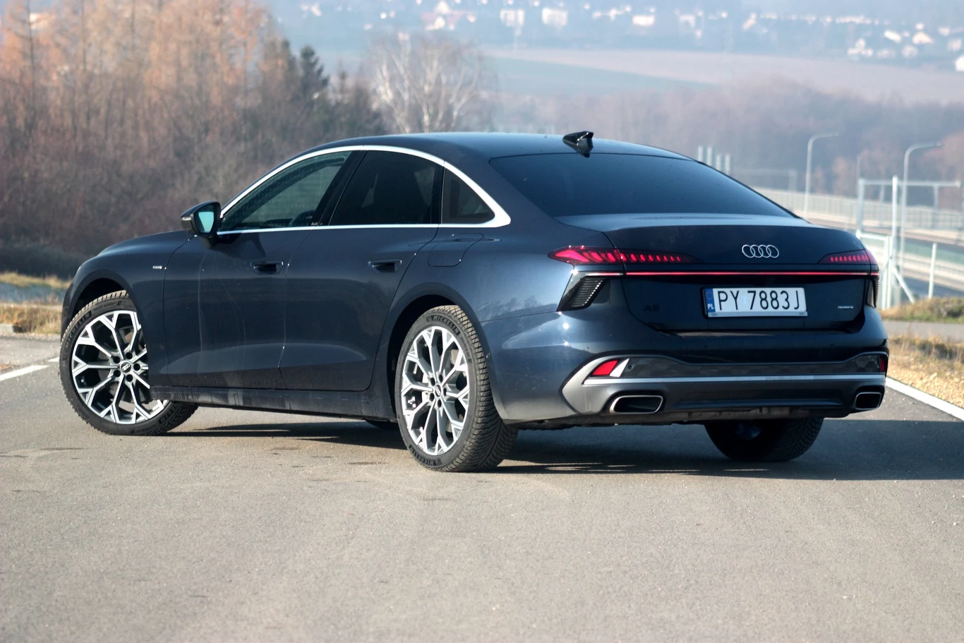 Granatowe Audi na szerokich, aluminiowych felgach stoi na pustej drodze, tyłem w stronę obiektywu, z wyraźnie widocznym logo marki oraz eleganckimi, przyciemnionymi tylnymi szybami i światłami LED.