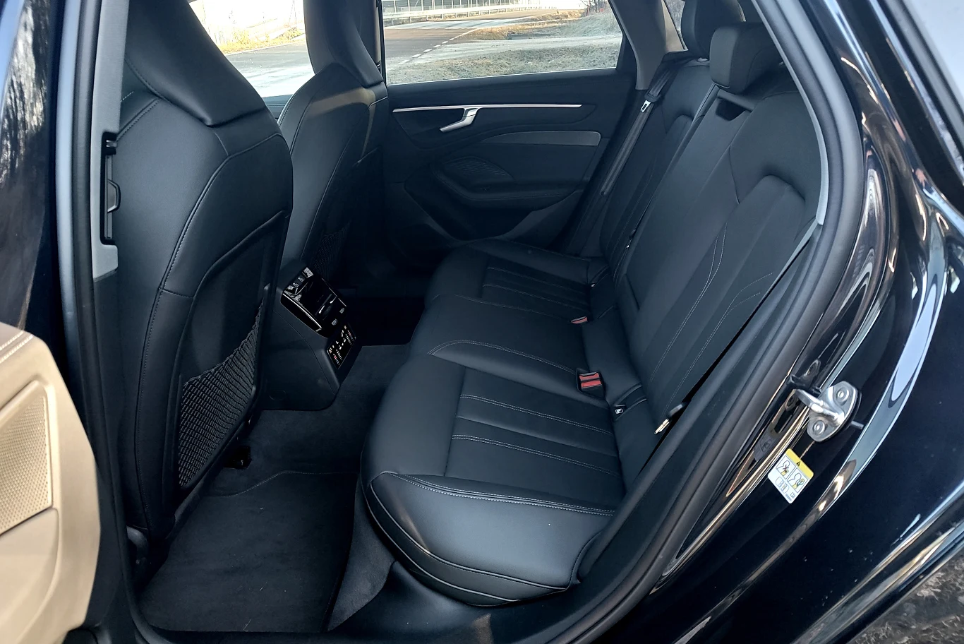 Czarne, skórzane tylne siedzenia samochodu z widocznymi pasami bezpieczeństwa, środkowym podłokietnikiem i nawiewami powietrza przy tylnej części konsoli; wnętrze przestronne i dobrze doświetlone przez otwarte drzwi.