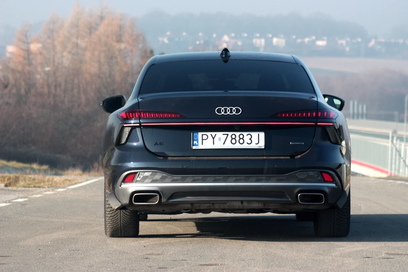 Czarny samochód Audi A6 zaparkowany na asfaltowej drodze, widoczny od tyłu z charakterystycznymi światłami LED i podwójnymi końcówkami wydechu, otoczenie stanowi rozmyty krajobraz z zaroślami i wzgórzami w tle.