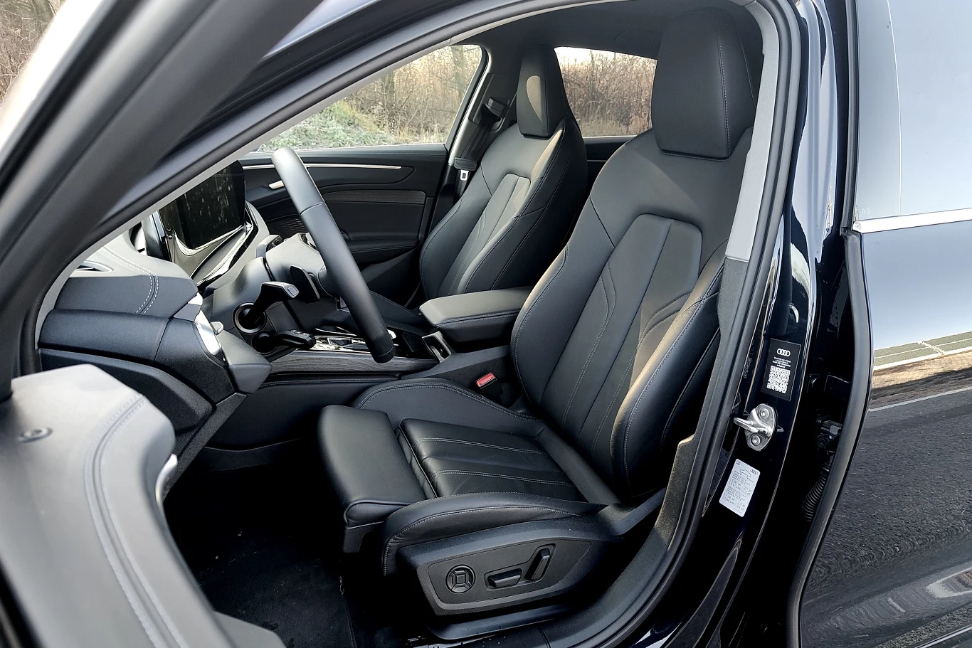 Wnętrze nowoczesnego samochodu z dwoma czarnymi, skórzanymi fotelami, przejrzystą deską rozdzielczą oraz sportową kierownicą; widoczne elektryczne sterowanie siedzeń i duży ekran dotykowy na konsoli środkowej.