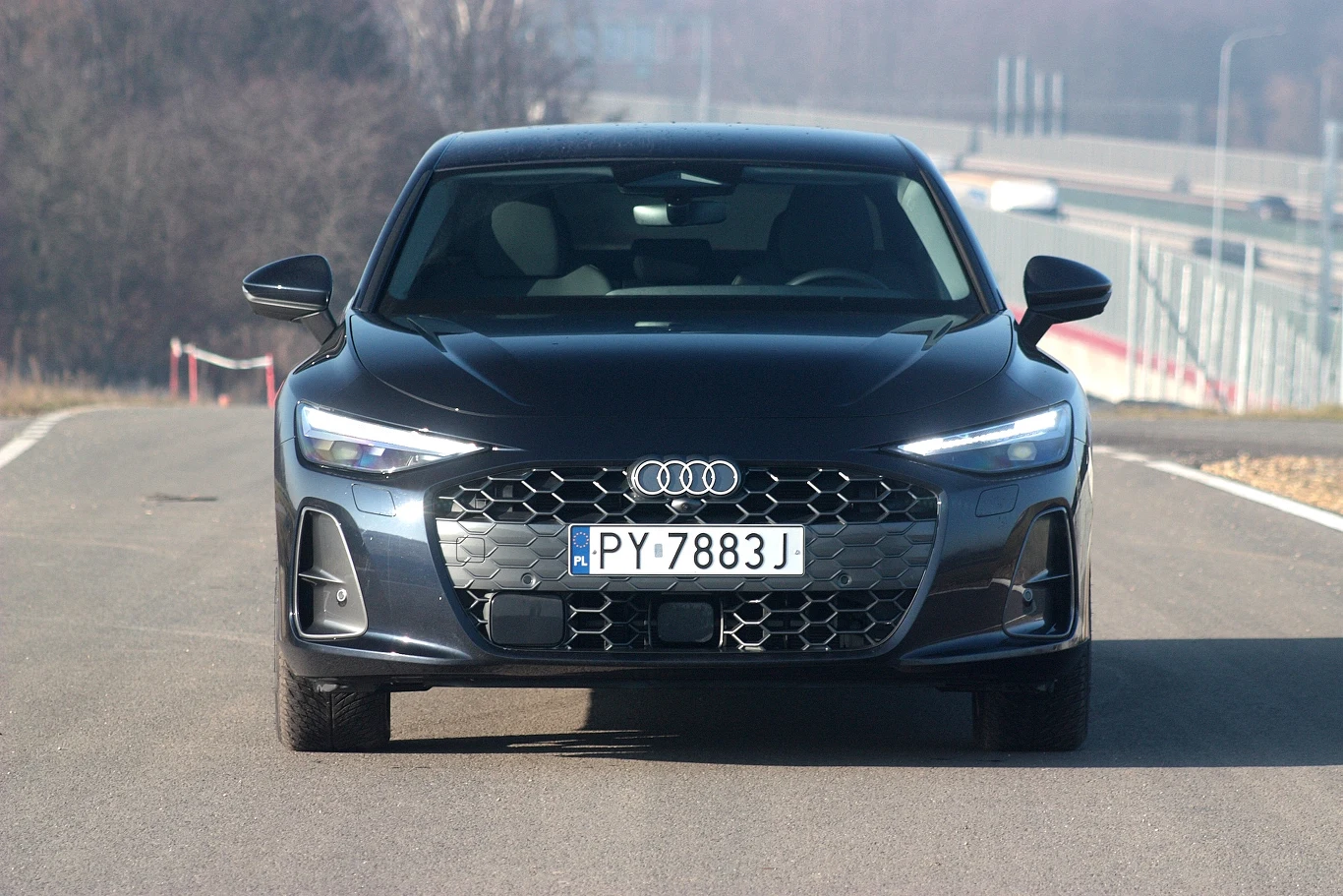 Czarny samochód marki Audi o nowoczesnym wyglądzie, z szeroką atrapą chłodnicy, charakterystycznymi światłami LED i polską tablicą rejestracyjną PY7888J, ustawiony centralnie na pustym fragmencie drogi.