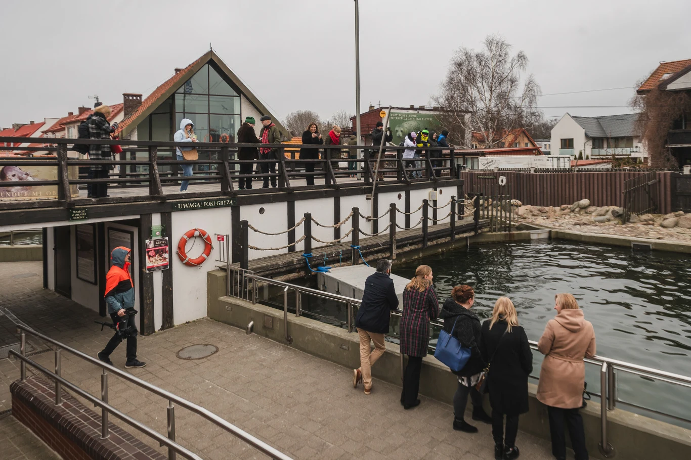 Grupa osób obserwuje wybieg dla zwierząt z wodnym basenem, wokół którego stoją zabezpieczenia i barierki, w tle charakterystyczna zabudowa oraz drzewa bez liści.