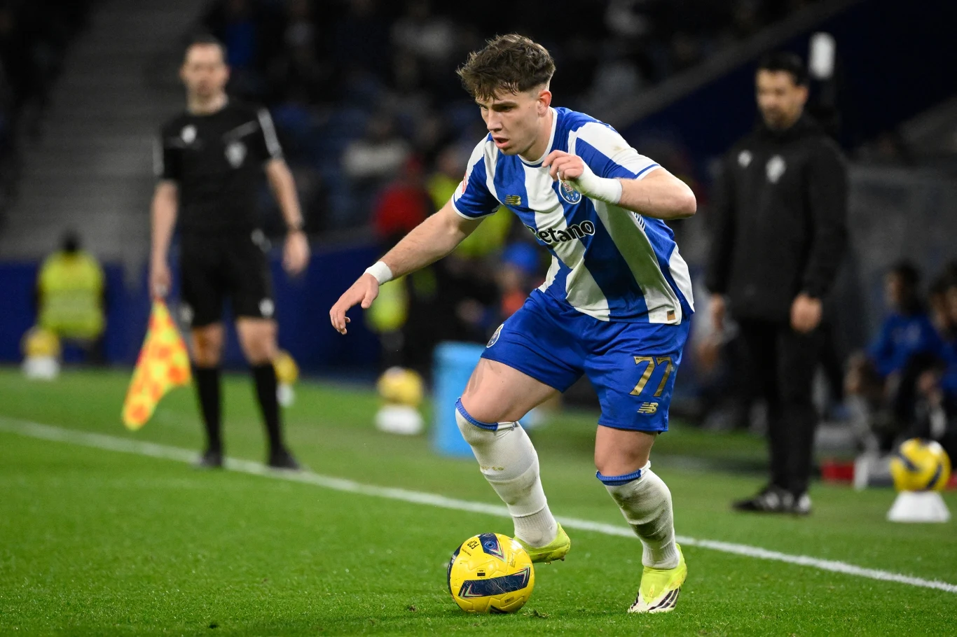 Piłkarz w niebiesko-białym stroju prowadzi piłkę przy linii bocznej boiska podczas meczu piłkarskiego, a w tle widoczni są sędziowie i osoby z drużyny przy linii bocznej.