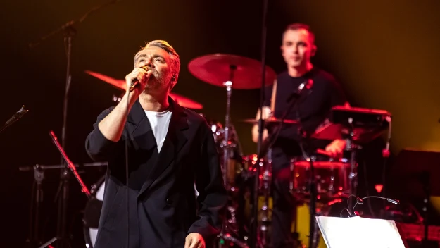 Wokalista w eleganckim, ciemnym garniturze śpiewa do mikrofonu na scenie koncertowej, w tle perkusista za zestawem perkusyjnym, z przodu widoczny pulpit z nutami, całość oświetlona kolorowymi światłami scenicznymi.