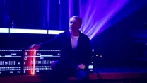 Mężczyzna w średnim wieku klęczący na scenie, ubrany w ciemny strój, na tle podświetlonych na fioletowo elementów dekoracji oraz elektronicznego panela z napisami i numerami, otoczony teatralnym oświetleniem.