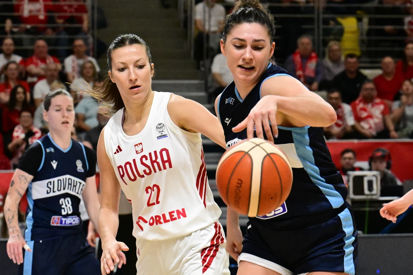 Zawodniczka drużyny Polski w białym stroju i koszulce z numerem 22 rywalizuje o piłkę z koszykarką drużyny Słowacji podczas dynamicznej akcji na parkiecie; w tle widoczni są kibice i pozostałe zawodniczki.