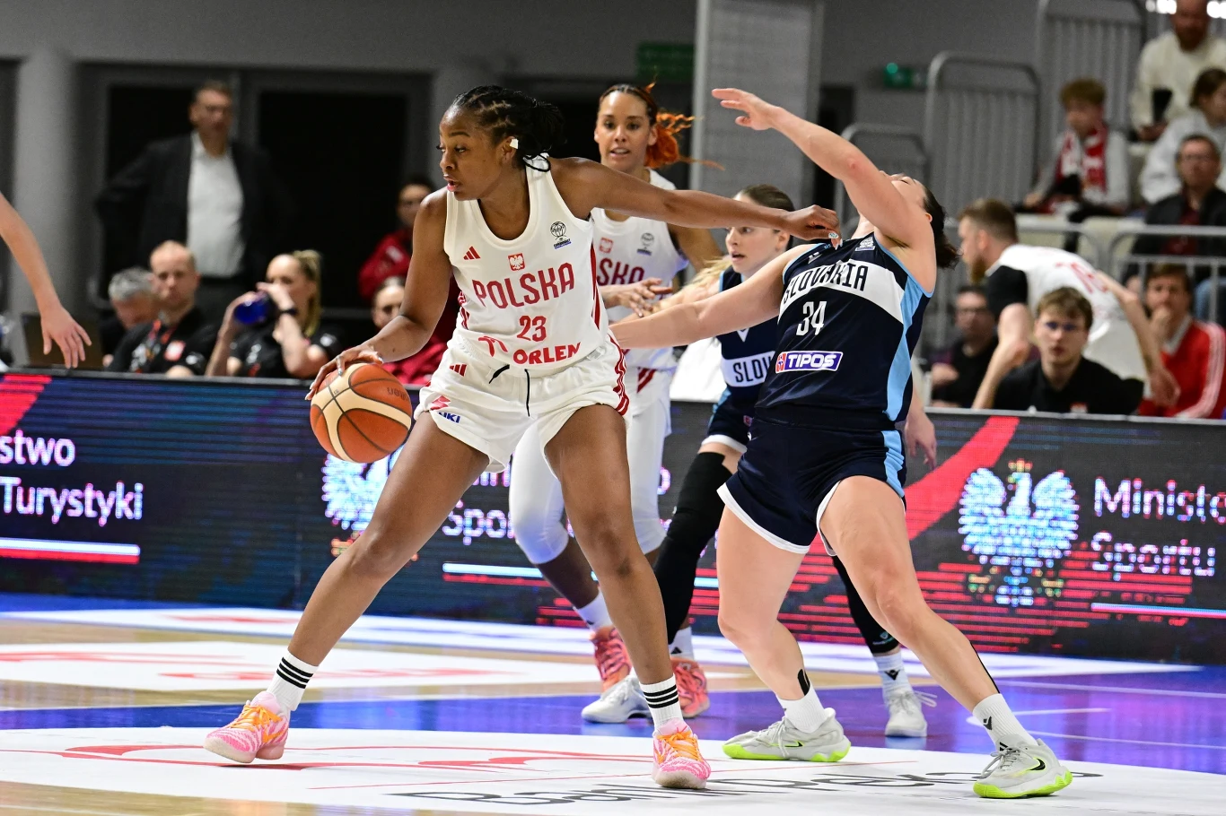 Zawodniczka drużyny Polski w białym stroju z numerem 23 próbuje przedrzeć się przez obronę rywalki ze Słowacji w granatowym stroju na parkiecie hali sportowej podczas meczu koszykówki, z piłką w dłoni oraz obecnością sędziów i kibiców w tle.
